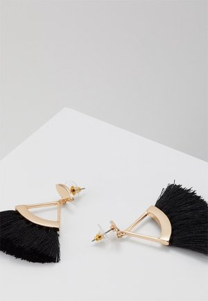 Boucles d'oreilles en métal doré avec des pampilles en franges noires posées sur une surface blanche, conçues sous forme de formes géométriques avec attaches à tiges.