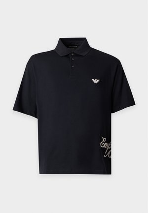 Homme debout portant un polo noir à manches courtes avec logo et inscription, pantalon large noir et chaussures noires sur un fond uni.