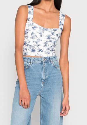 Vrouw draagt een wit cropped topje met blauwe bloemenprint en lichtblauwe hoge taille jeans, staand tegen een effen achtergrond.