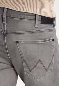 Grå denimjeans med en bakficka med en zickzack-sticksdesign och en svart läderpatch på midjan. Texturerad, åtsittande design.