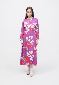 Μωβ φόρεμα μακρυμάνικο με λουλουδάτο μοτίβο σε κόκκινο και λευκό, με εφαρμοστή μέση και ρέουσα σιλουέτα. Φορεμένο με ανοιχτόχρωμα παπούτσια.