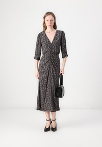 Langes, dunkles Kleid mit V-Ausschnitt, Dreiviertelärmel, geraffter Taille und einem bunten abstrakten Muster. Kombiniert mit einer kleinen schwarzen Handtasche.