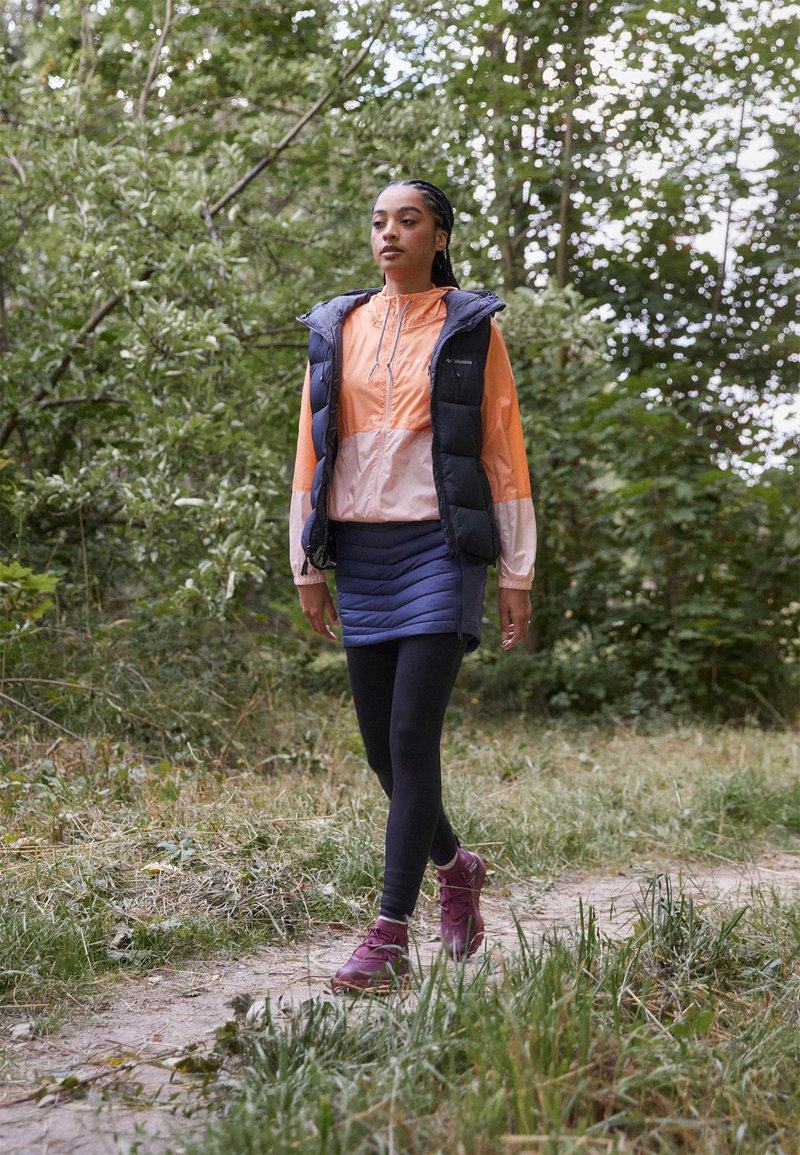 Odzież na zewnątrz składająca się z czarnej kamizelki puchowej, pomarańczowej i różowej kurtki z zamkiem, granatowej spódnicy, czarnych legginsów oraz fioletowych butów trekkingowych, osadzona w lesie.