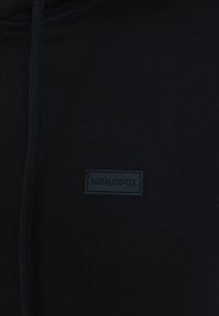 Hoodie noir avec une texture de tissu lisse, présentant un cordon de serrage et une étiquette rectangulaire noire sur laquelle est inscrit « SMILODOX ». 