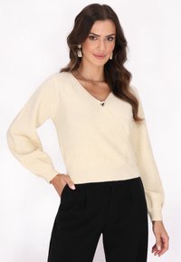 Heller beigefarbener Wickelpullover aus weichem Material, mit V-Ausschnitt und puffartigen langen Ärmeln, kombiniert mit schwarzen hochgeschnittenen Hosen.