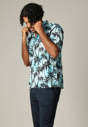 Chemise à manches courtes à boutons de couleur bleu clair avec un imprimé de palmiers noirs, dotée d'un col et d'une coupe décontractée, portée avec un pantalon sombre.