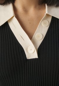 Haut noir côtelé avec un col couleur crème et trois boutons crème légèrement brillants. Le tissu présente une texture verticale et des coutures subtiles.