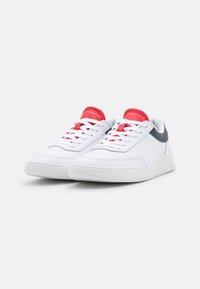 Sneaker bianche con accenti rossi sulla linguetta e i lacci, con un controfibbie grigia e dettagli perforati sui lati. Suola in gomma.