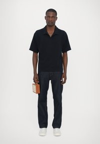 Tricou polo negru cu mânecă scurtă, blugi de denim albastru închis, adidași albi și o geantă rectangulară maro cu un detaliu de închidere aurie.