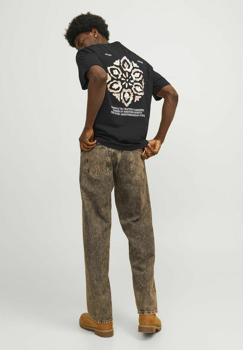 Zwarte t-shirt met korte mouwen met een kleurrafloraal grafisch ontwerp op de achterkant. Gekleed met vervaagde, loszittende bruine spijkerbroek en bruine schoenen.
