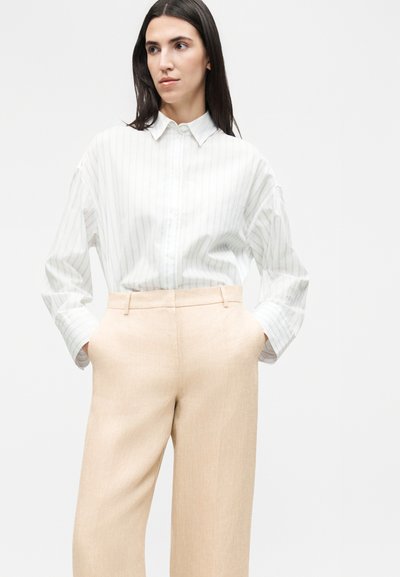 Γυναίκα με μακριά σκούρα μαλλιά που φοράει μια λευκή ριγέ πουκάμισο με κουμπιά μαζεμένο μέσα σε μπεζ ψηλόμεσο παντελόνι με τα χέρια στις τσέπες.