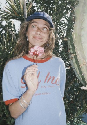 Niebieska koszula z czerwonymi akcentami, na której czerwonym kolorem nadrukowano napis "Surf Goods", trzyma różowy kwiat, nosi srebrne bransoletki i niebieską czapkę, otoczony zielenią.