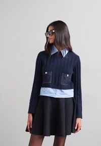 Suéter recortado de punto acanalado azul marino con dos bolsillos de denim en el frente, superpuesto a una camisa de color azul claro con cuello, combinado con una falda negra acampanada.