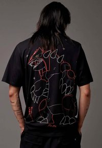 Schwarzes T-Shirt mit einem großen roten und weißen Grafik eines Wesens mit scharfen Krallen und Stacheln, auf dem Rücken gedruckt. Kurze Ärmel, lässige Passform.