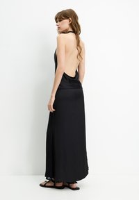 Black satin dress with an open back design, flowing silhouette, and side split, complemented by flat sandals and loose, wavy hair.