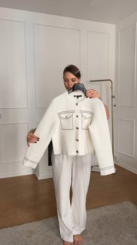 Giacca bianca a cropped con cuciture nere, dotata di due tasche frontali e bottoni argentati, appesa a un appendiabiti su uno sfondo neutro.