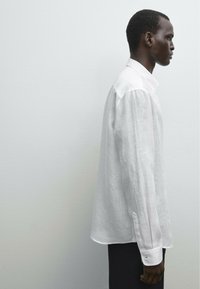 White linen button-up shirt with a pointed collar, long sleeves, and cuffs. Loose fit and a smooth texture, displayed against a neutral background.