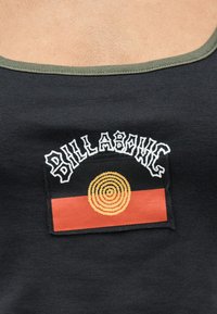 Μαύρο αμάνικο τοπ με πράσινο περίγραμμα που φέρει ένα λάβαρο με την επιγραφή «Billabong» πάνω από ένα κόκκινο και μαύρο φόντο με κίτρινο σχεδιασμό με ομόκεντρους κύκλους.