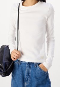 Vit ribbad långärmad tröja med rund halsringning och en liten etikett. Kombinerad med lösa blå denimshorts, där en hand håller en svart väska.