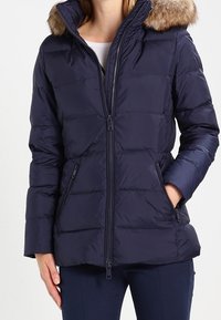 Donna che indossa un giubbotto invernale trapuntato di colore blu navy, con cappuccio bordato in pelliccia e chiusura zip, tasche con le mani in tasca.