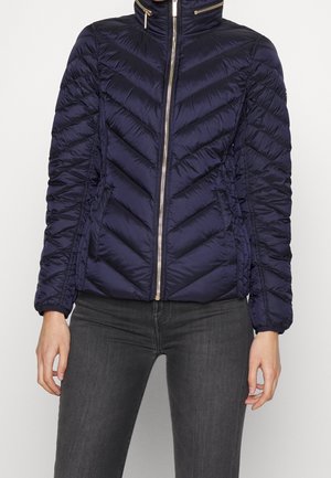 Personne portant une veste matelassée bleu marine à fermeture éclair avec des détails zippés dorés et un pantalon cintré gris foncé, sur un fond uni.