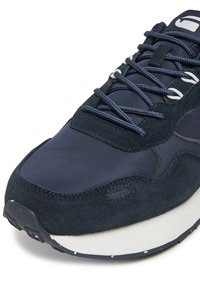 Tumši zils sporta apavs ar gludu sintētiskā materiāla un teksturēta zamša sajaukumu. Aprīkots ar apaļām šņorēm, mīkstinātu zolīti un baltām detaļām.