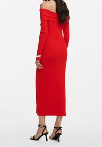 Robe rouge côtelée, épaules dénudées, manches longues, longueur mi-mollet, fente sur le côté, portée avec des talons noirs à bride cheville.