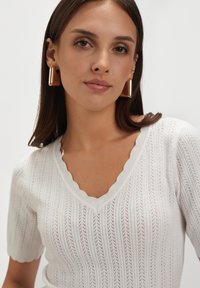 Biała dzianinowa bluzka z dekoltem w serek i falbaniastymi brzegami. Posiada prążkowaną teksturę z otwartymi wzorami. Noszona z złotymi prostokątnymi kolczykami.