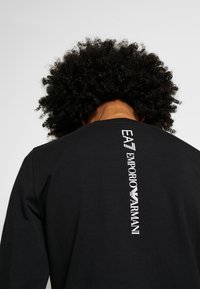 Svart långärmad T-shirt med vertikal vit logotyptext på ryggen. Tillverkad av mjukt material med en slät yta.