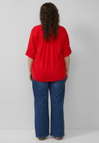 Rote Kurzarmbluse mit umgeschlagenen Ärmeln, kombiniert mit weit geschnittenen blauen Jeans. Die Bluse hat einen gerafften Rücken für zusätzliches Volumen.