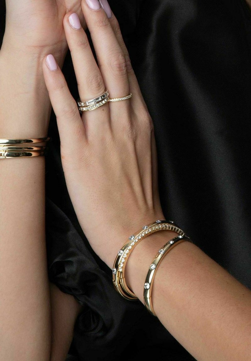 Guldtonade metallarmband med klara rhinestones, som har olika former och mönster, bärs på händer med ljusrosa nagellack.