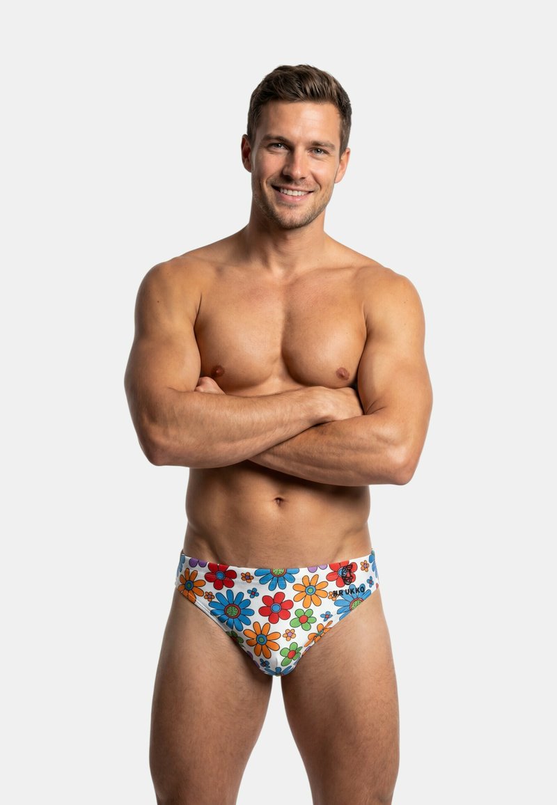 Lachende gespierde man met gekruiste armen, gekleed in witte zwembroek met kleurrijke bloemenpatronen, staand tegen een effen achtergrond.