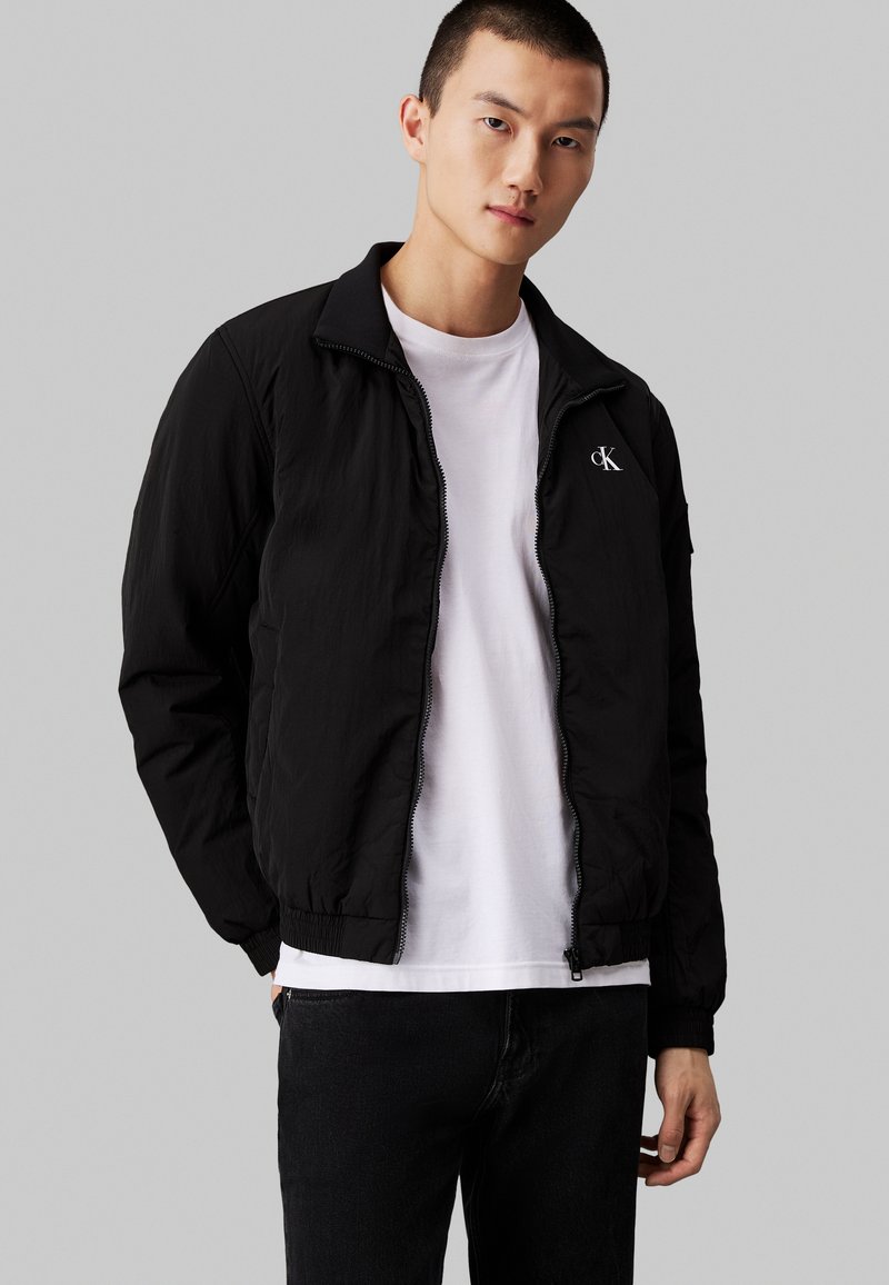 Melnā zip-up bomber jaka ar stāvu apkakli, struktūras audumu un elastīgām piedurknēm. Ar baltu Calvin Klein logo uz krūtīm.