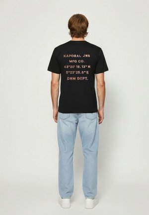 Homme portant un T-shirt noir avec des coordonnées imprimées et du texte, un jean bleu clair et des baskets blanches, debout de dos sur un fond uni.