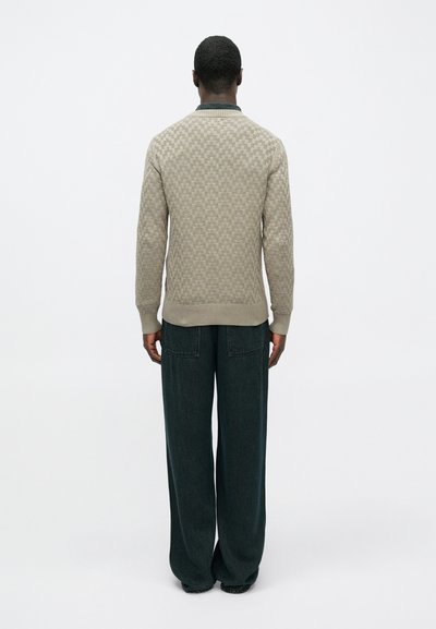 Vīrietis stāv ar muguru pret skatītāju, ģērbies beigē teksturētā džemperī un brīvās tumši zilās biksēs, uz fona ar vienkāršu baltu krāsu.