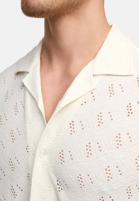 Camicia a maniche corte color crema con motivo a occhiello, tessuto testurizzato, colletto a risvolto e chiusura con un solo bottone al collo.