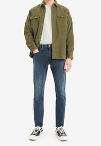 Green corduroy shirt, gray T-shirt, blue jeans, black and white sneakers; front view; two chest pockets on the shirt.