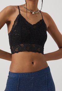 Brassière en dentelle noire avec des bords festonnés et des fines bretelles ajustables. Le tissu présente des motifs floraux et un design court, mettant en valeur la texture.