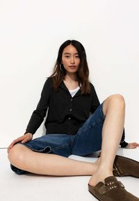Cardigan noir à boutons, short en denim bleu avec ourlet effiloché, chaussures brunes à enfiler avec clous métalliques, pose assise sur fond blanc.