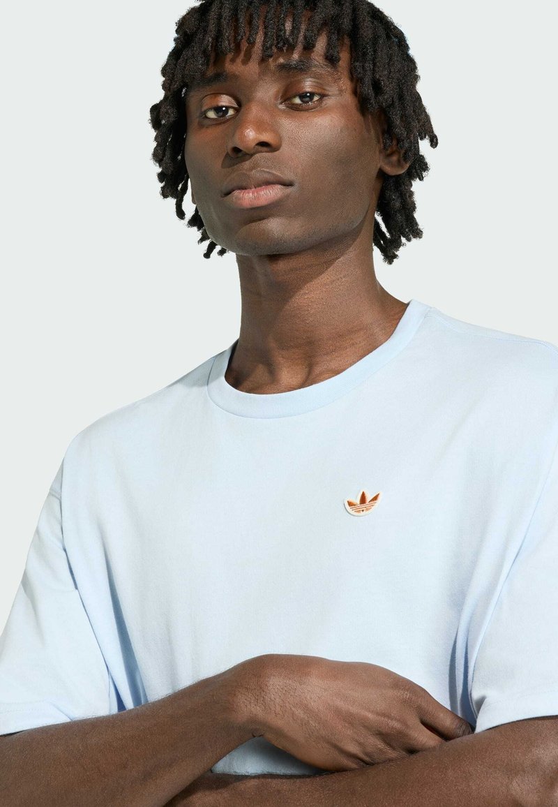Ung sort mand med korte dreadlocks iført en lyseblå Adidas t-shirt med korslagte arme mod en ensfarvet baggrund.