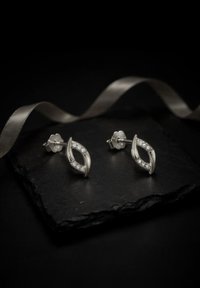 Boucles d'oreilles en argent en forme d'œil, ornées de petits cristaux ronds transparents intégrés au centre, présentées sur un fond ardoise foncé.
