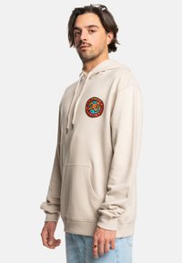 Béžová mikina s kapucňou a prednými vreckom, s farebným kruhovým logom na hrudi. Mäkký materiál a voľný strih.