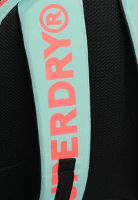 Primer plano de una correa de mochila color teal con el texto coral "SUPERDRY®" sobre un fondo de malla negra.