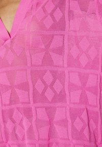 Rosa stickad tyg med en diamantskiss och olika strukturerade rutor. V-ringad design med en lätt och andningsbar textur.