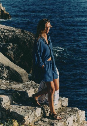 Woman in blue shirt and shorts walks barefoot on rocky shore with ocean in the background, holding a striped towel.