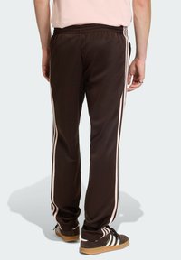 Brązowe spodnie dresowe z białymi paskami po bokach, wykonane z gładkiego materiału. Posiadają elastyczny pas i zwężane mankiety. Wyróżniają się sportowym stylem.