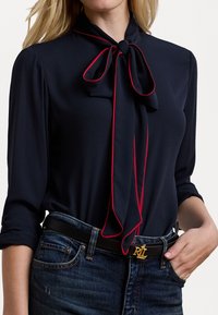 Bluză bleu marin cu un accent roșu legat, din material moale, cu mâneci lungi și manșete răsucite, asortată cu blugi negri și o curea neagră cu accesorii din aur.