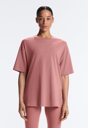 Frau mit zurückgebundenem dunklem Haar, die ein lockeres, staubiges rosa Kurzarmoberteil und passende Hose trägt, stehend vor einem schlichten hellen Hintergrund.