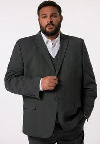 Completo grigio in tessuto texture, giacca monopetto con rever a norma e una leggera lucentezza. Abbinato a una camicia bianca e a un gilet grigio.