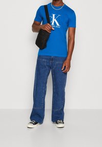 Modrá bavlněná trička s bílým logem, kombinovaná s širokými denimovými džíny. Černá kabelka přes rameno a černé tenisky s bílými akcenty.